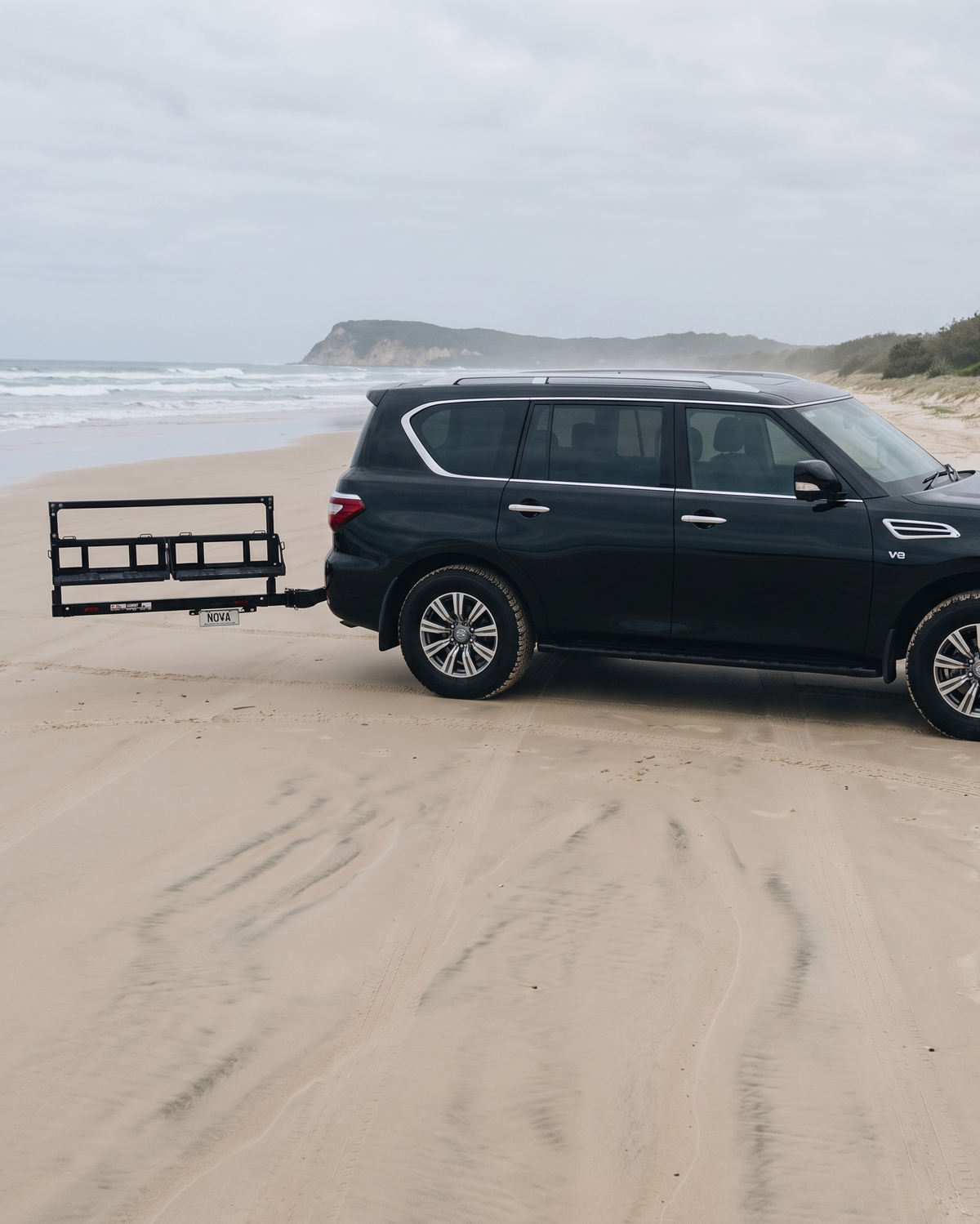 A black SUV parked on a sandy beach with the ocean and dunes in the background, towing a black utility trailer.