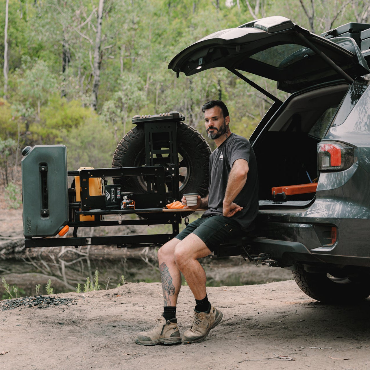 Man sitting by an open car trunk with a trailer attached in a forest setting